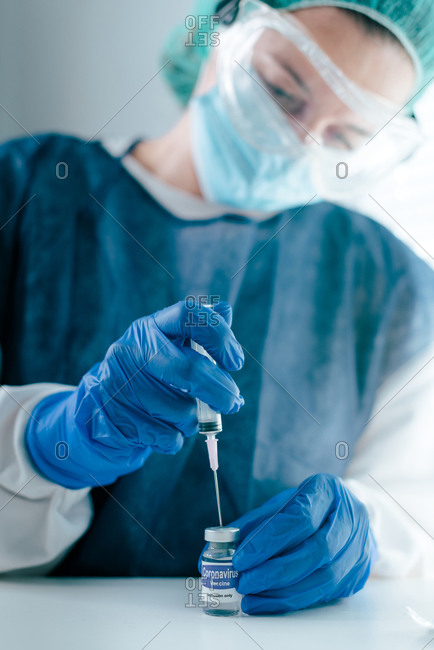 Young female medical specialist in protective mask and gloves filing syringe with vaccine from vial for prevention and immunization from coronavirus infection