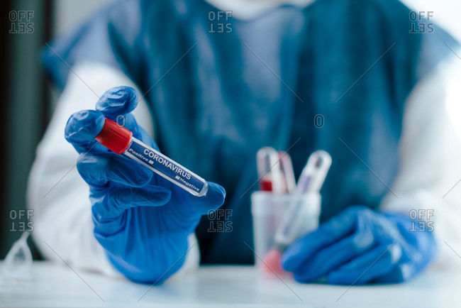 Crop anonymous medical worker in protective uniform and latex gloves holding test tube with blood sample for coronavirus test while working in lab