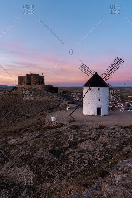 Aged windmill located on rocky cliff near medieval castle against cloudy sundown sky in countryside