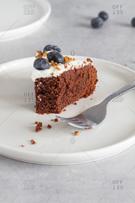 Slice of homemade chocolate sponge cake topped with fresh whipped cream and whole blueberries placed on table in light kitchen
