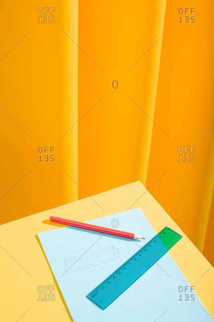 Red pencil and blue ruler with papers and draws over a yellow table