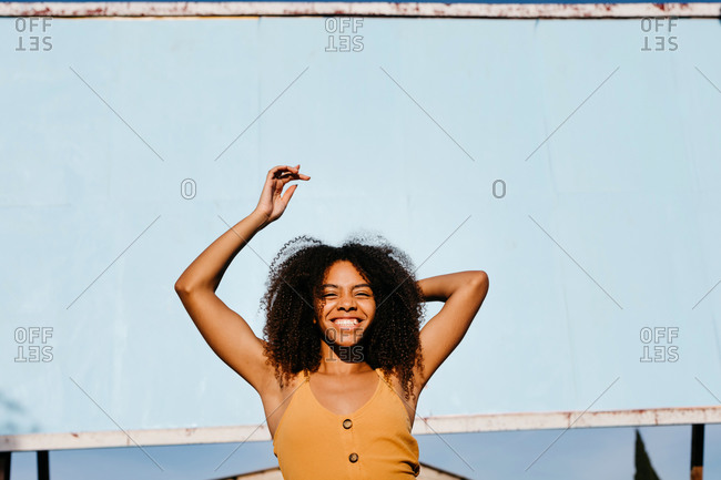 Low angle of happy African American female in brown summer dress smiling at camera while standing with arms raised on street against blank billboard in sunlight