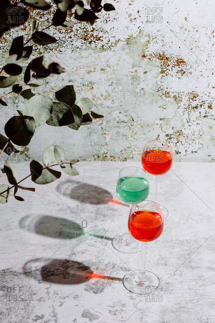From above of glasses filled with colorful liquids put on concrete surface near plant with dark green leaves and rusty wall