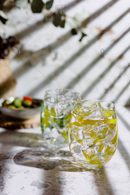 Large glasses of mojito cocktail with mint leaves and ice cubes put on concrete surface near plate with gherkins and vase with plant near shabby wall