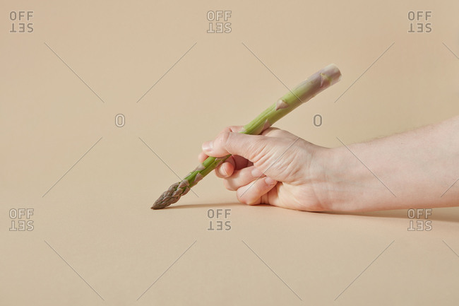 Woman hand is painting by fresh natural organic asparagus spear as a paining brush on a sand yellow background, copy space.