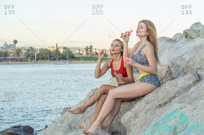 Full body barefoot women in swimwear sitting on stones and blowing bubbles near sea in evening on resort