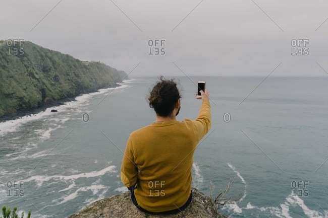 Rear view of man sitting on a rock at the coast taking a smartphone picture on Sao Miguel Island- Azores- Portugal