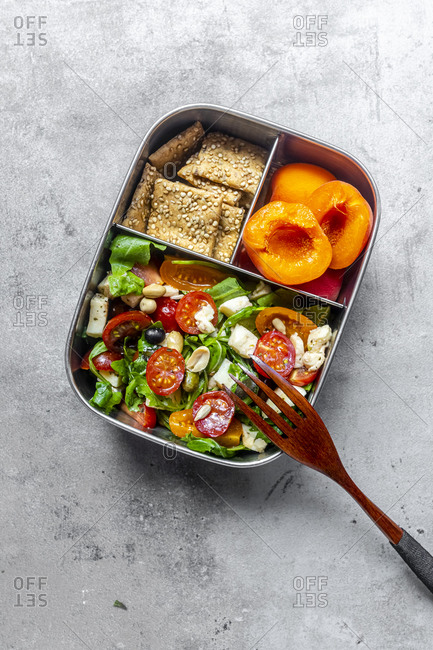 Lunch box with rocket salad with colored tomatoes- mozzarella and nuts- crispbread and apricots- wooden fork on concrete surface