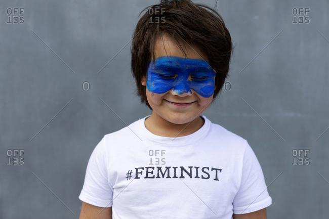 Portrait of smiling boy with painted blue mask on his face wearing t-shirt with imprint 'Feminist'