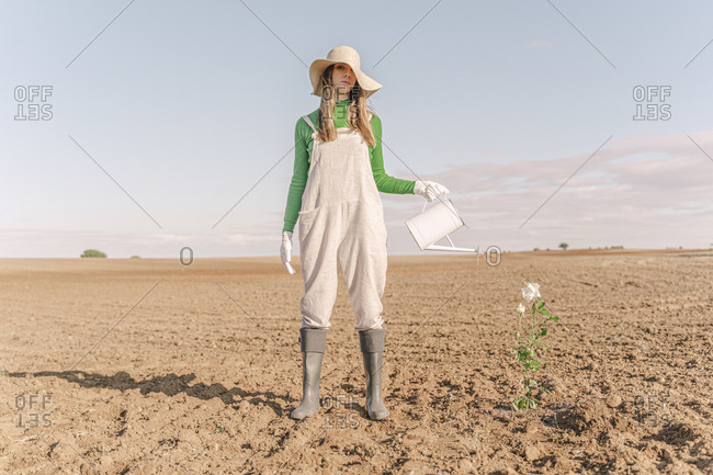 Woman standing on barren field- watering flower