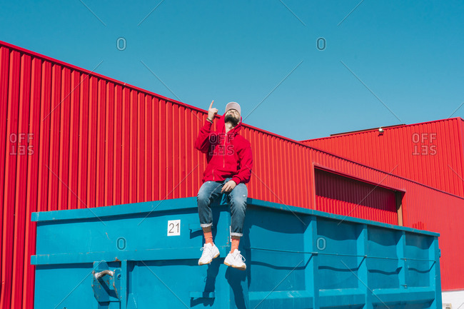 Young man wearing red hoodie- sitting on edge of blue container in front of red wall- raising forefinger
