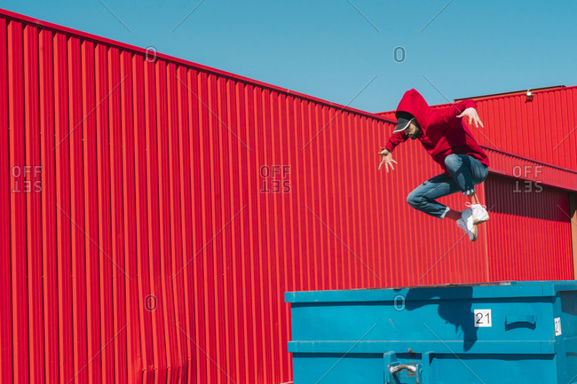 Young man wearing red hooded jacket jumping from edge of container in front of red wall