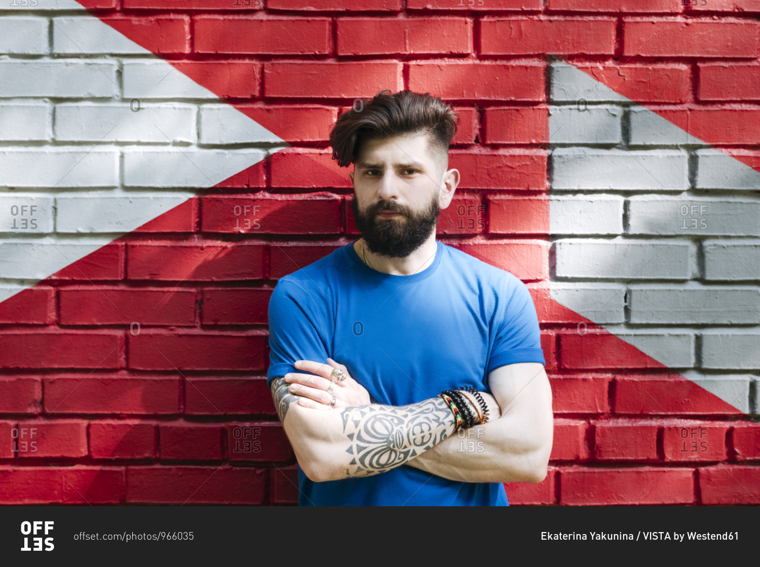 Young man with tattoo standing in front of red brick wall with arms crossed