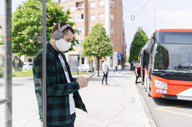 Young man with headphones wearing protective mask using smartphone while waiting at bus stop- Spain