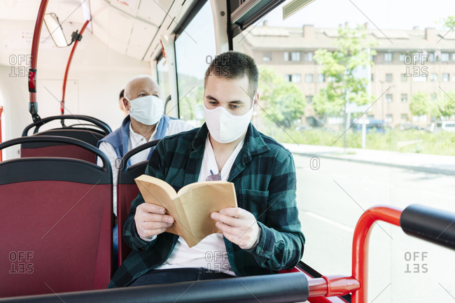 Portrait of young man wearing protective mask and gloves in public bus reading a book- Spain