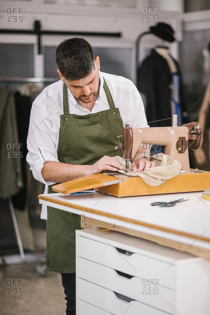 Diligent focused male tailor in apron sewing outfit details using modern sewing machine at table while creating exclusive clothes collection in contemporary work studio