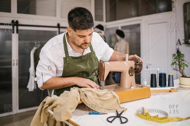 Diligent focused male tailor in apron sewing outfit details using modern sewing machine at table while creating exclusive clothes collection in contemporary work studio