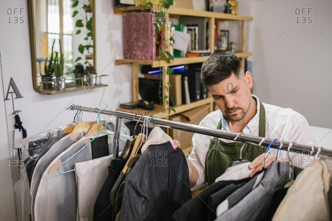 High angle of serious focused male tailor in apron checking details of apparel hanging on hanger on metal rack among other trendy bespoke clothes in modern workroom