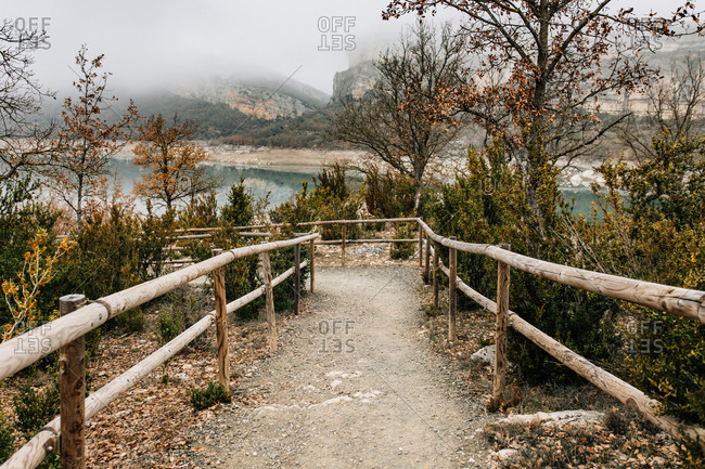 Empty curved path with wooden railing leading among trees with dry foliage on hill slope near mountain lake in foggy day in Montsec Range in Spain