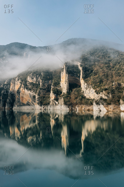 Magnificent landscape of calm lake with mirrored water surface surrounded by rough rocky mountains of Montsec Range covered with dense fog in cold day in Spain