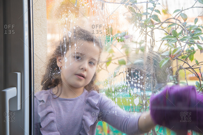 Positive little girl in casual dress with spray bottle of cleanse washing glass in room with rainbow painted on window during coronavirus quarantine