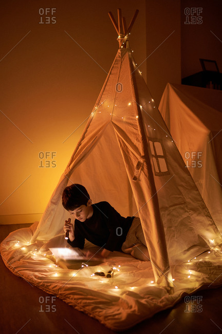 Focused little kid in casual wear reading book while lying in kids play tent with flashlight and garland around near fireplace in apartment