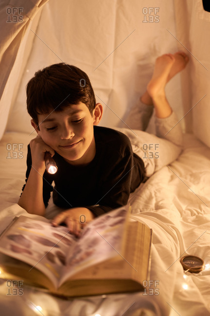 Focused little kid in casual wear reading book while lying in kids play tent with flashlight and garland around near fireplace in apartment