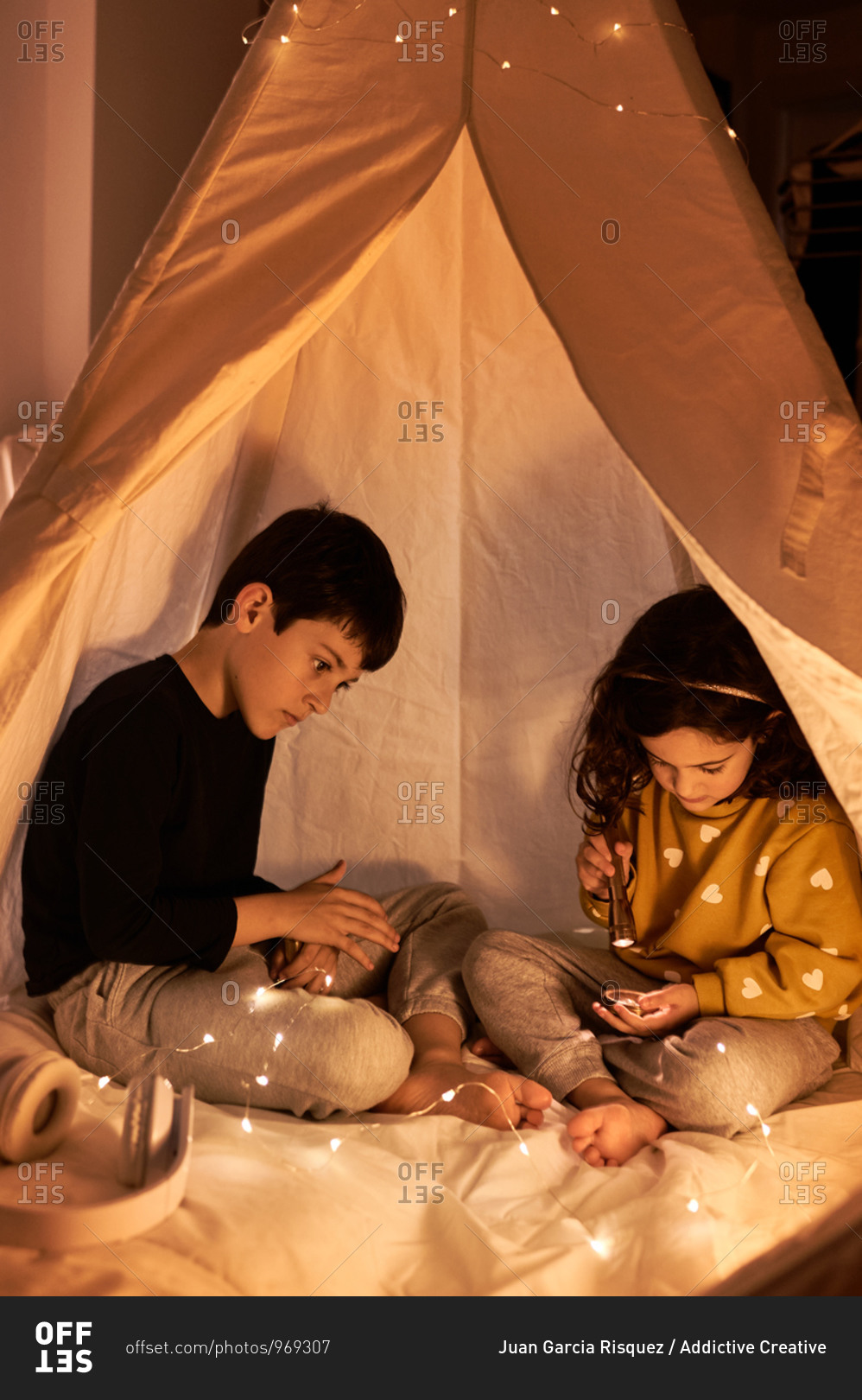 Curios boy watching sitting near dreamy little sister in casual wear playing with flashlight in garland tent in evening in flat