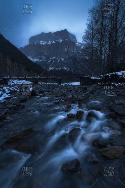 Narrow small creek in the middle of thin trees running through mountain valley covered partially with snow under cloudy sky at nightfall in wintertime