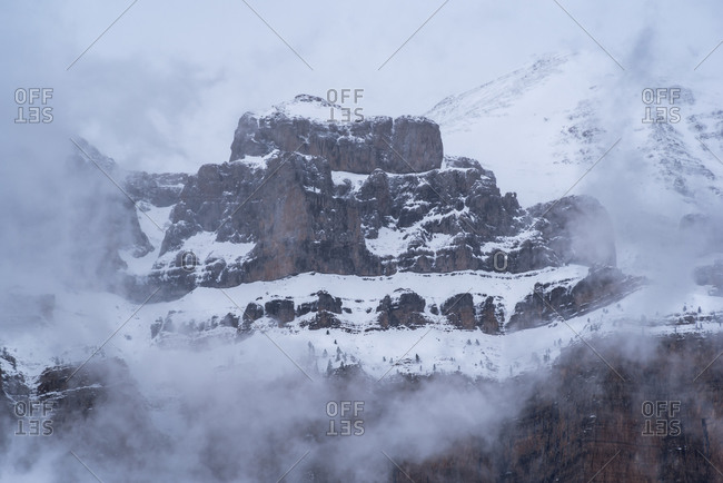 Sharp mountain peaks covered partially with snow surrounded by misty fog under cloudy sky in wintertime