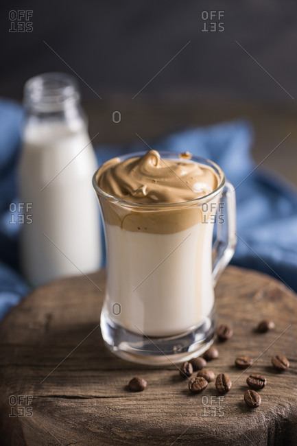 From above whipped coffee Dalgona coffee in cold milk placed near coffee beans and milk bottle on rustic wooden round board surface with blue napkin underneath