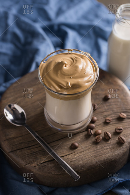 From above whipped coffee Dalgona coffee in cold milk placed near coffee beans, teaspoon and milk bottle on rustic wooden round board surface with blue napkin underneath