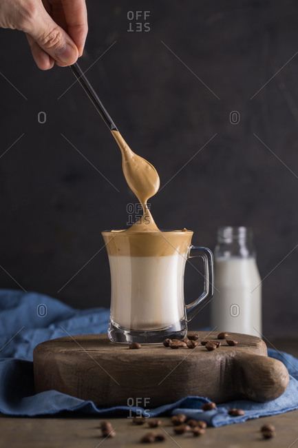 Cropped person hand adding with teaspoon whipped coffee Dalgona coffee in cold milk placed near coffee beans, teaspoon and milk bottle on rustic wooden round board surface with blue napkin underneath