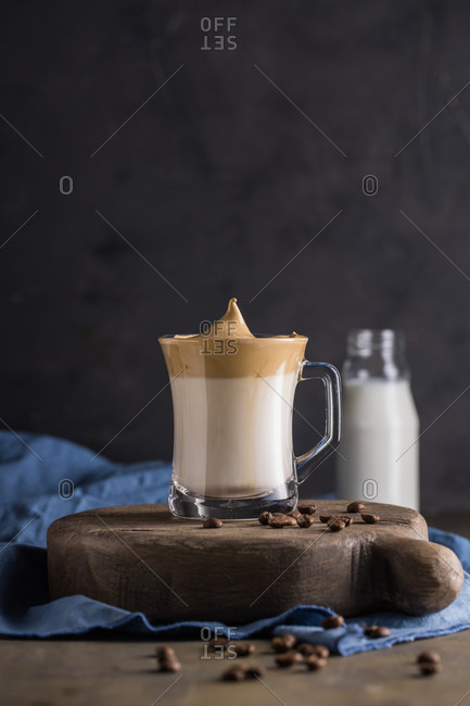 Whipped coffee Dalgona coffee in cold milk placed near coffee beans, teaspoon and milk bottle on rustic wooden round board surface with blue napkin underneath