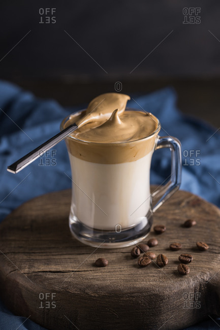 From above whipped coffee Dalgona coffee in cold milk placed near coffee beans and teaspoon on rustic wooden round board surface with blue napkin underneath