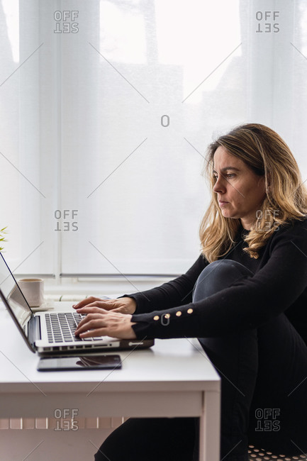 High angle side view of focused female specialist in casual wear working with electronic documents on laptop while sitting at table near window in modern room at home