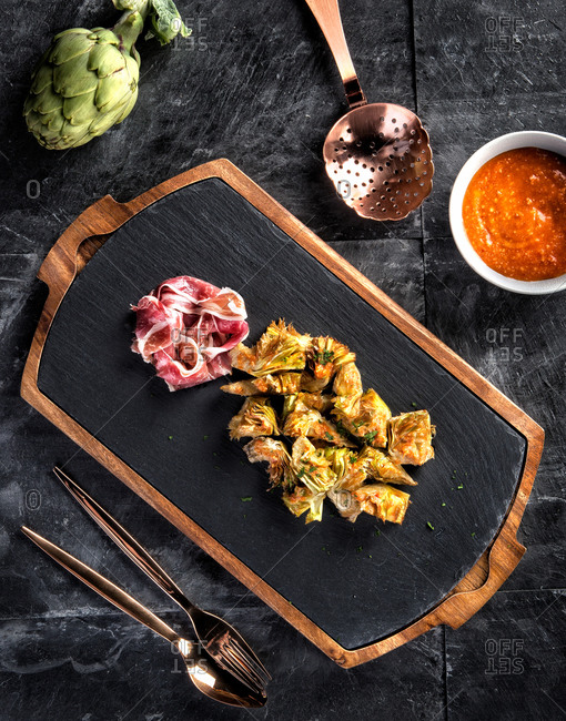 Top view composition with sliced ham and cooked artichokes served on black slate board on table with sauce and cutlery