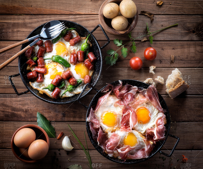 Pan with fried eggs with traditional Spanish iberian ham next to a pan with fried eggs, peppers and chorizo served on rustic wooden table with kitchen utensils and products