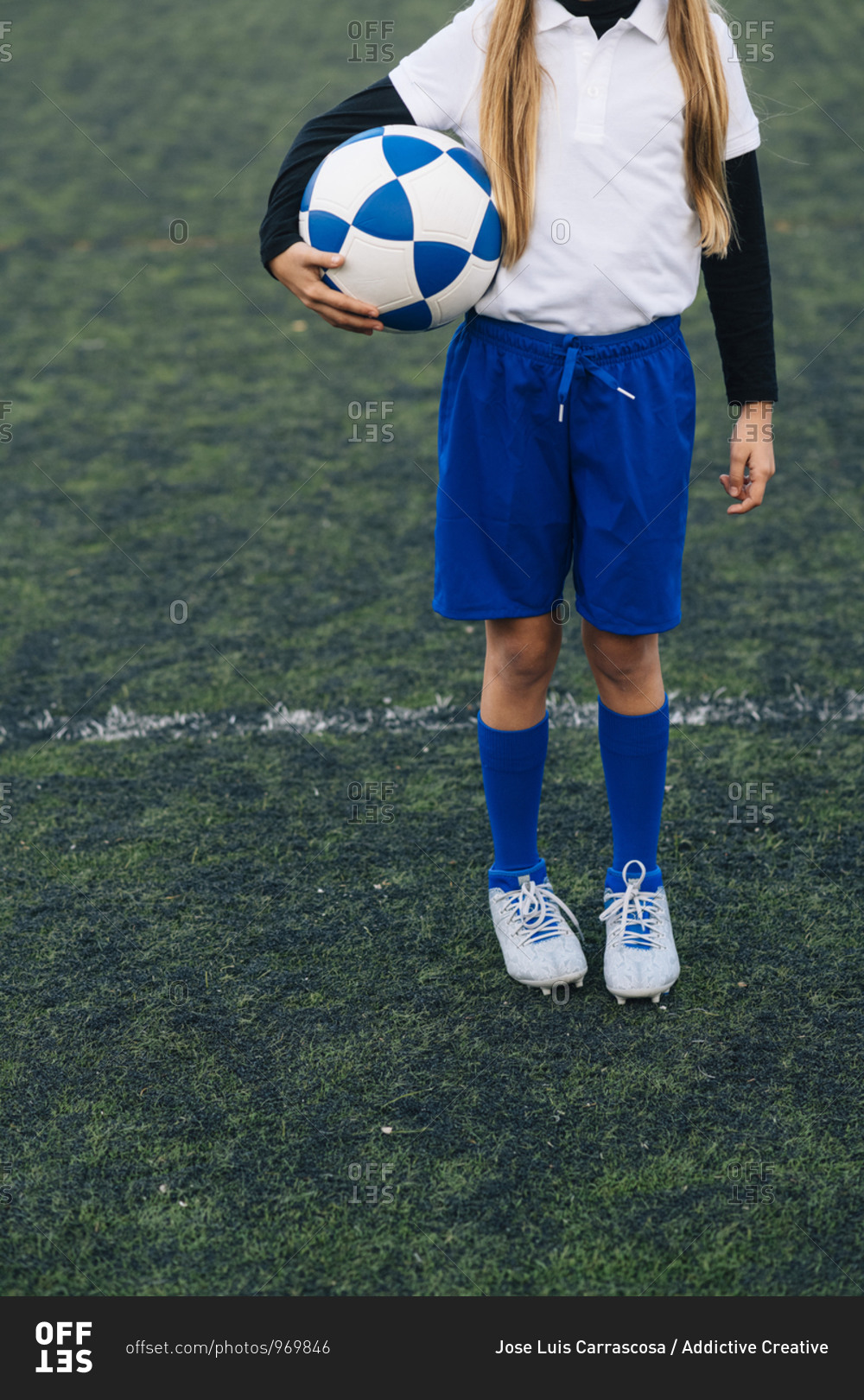 Crop girl in white and blue uniform with soccer ball while standing alone on green field in modern sports club