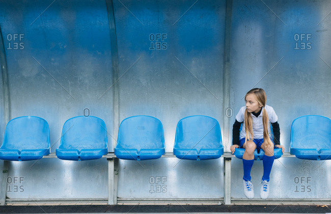 Full length frustrated preteen girl in soccer uniform sitting alone on blue plastic seat after match failure in sports club