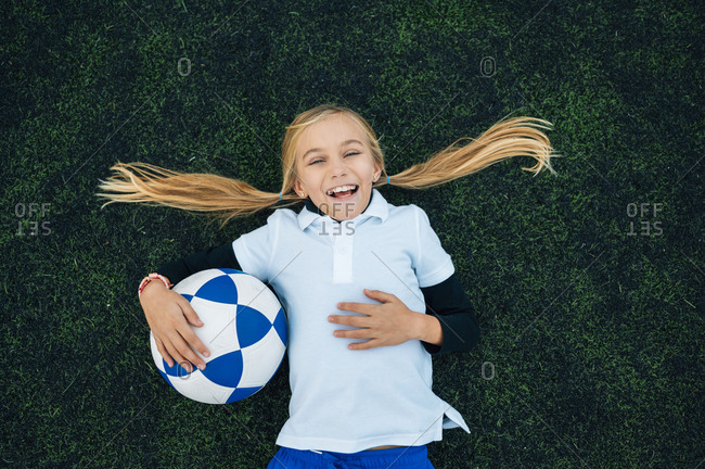 From above happy preteen girl with ponytails in uniform laughing while lying with ball on green field at football stadium