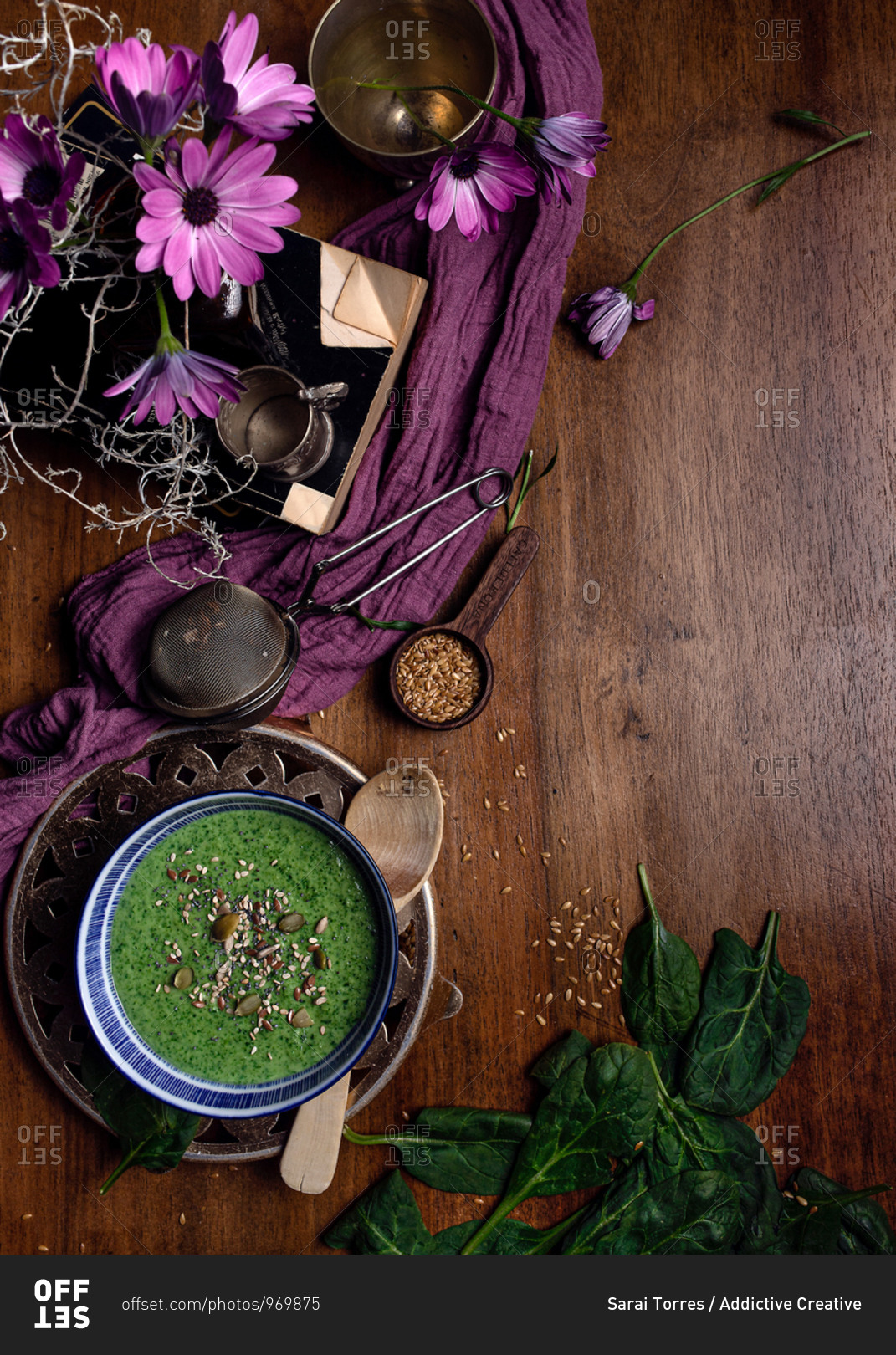 From above of ceramic bowl of fresh green smoothie with pumpkin seeds and sesame in composition with purple delicate fabric and bouquet of fresh violet Daisy flowers in vase placed on brown wooden table