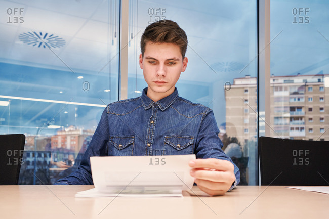Serious male worker in casual shirt sitting at table in modern open space workplace and reading documents