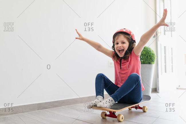 Little kid in red spotted helmet and casual clothes and dreaming about outdoors amusement while sitting alone on longboard in corridor of light modern apartment staying at home