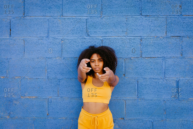 Calm African American female with Afro hairstyle and in yellow outfit leaning on blue brick wall in city and stretching arms at camera