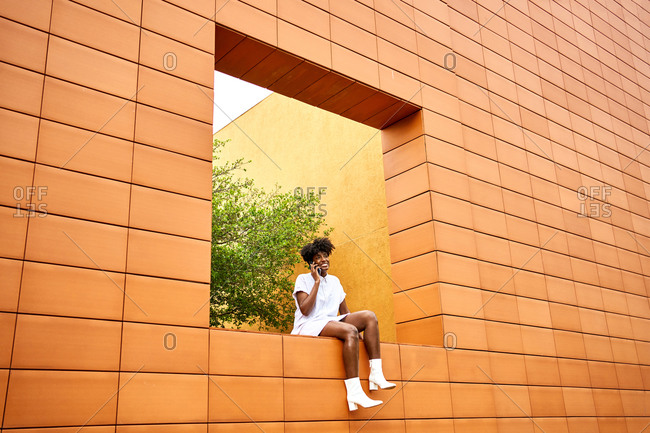 Low angle of young African American female in trendy white outfit smiling away and making phone call on street while sitting on wall contemporary building with bright orange and yellow walls