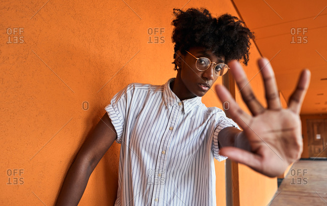 Adult African American female with eyes closed trying to stop negative impact while standing with arms raised against blurred vivid orange interior in corridor of modern building