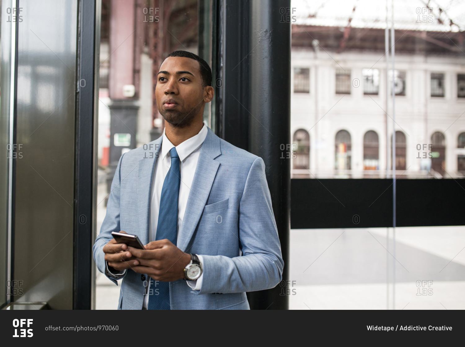 Serious black man in trendy suit holding smartphone and looking away thoughtfully through glass.
