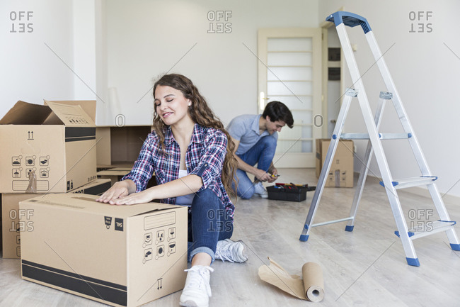 Young female using duct tape sitting near carton boxes and male sitting near black box in empty apartment