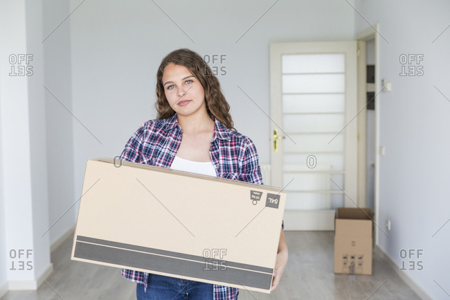 Young sweet female holding carton box in empty room looking at camera and smiling